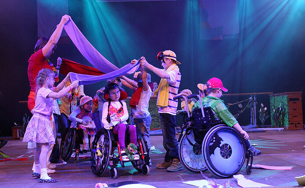 Eine Gruppe von Kindern steht in Kostümen auf einer Showbühne und tanzt mit schwingenden Tüchern. 2 Menschen im Rollstuhl sind auch dabeii.